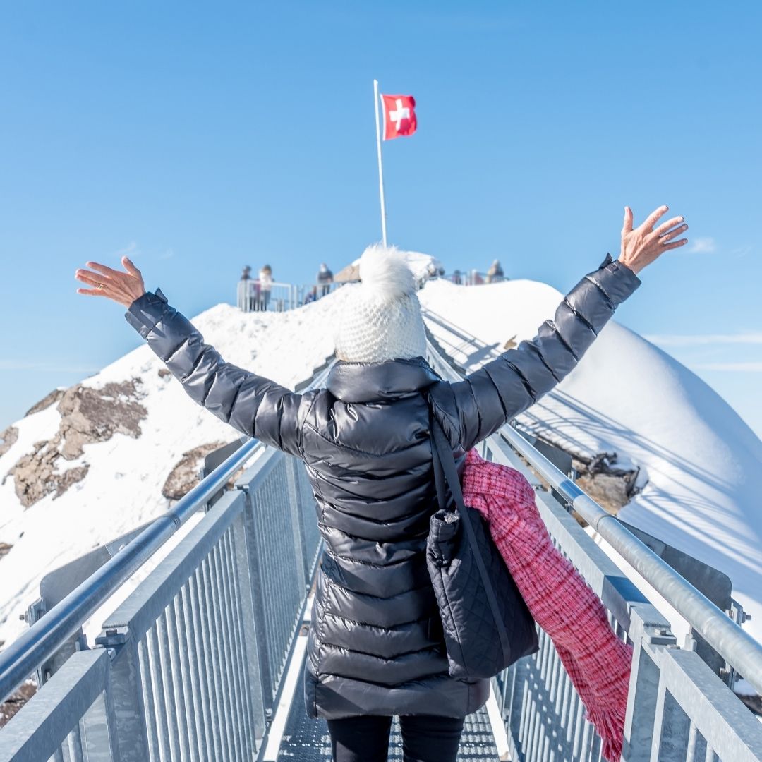 A traveller with arms raised on a high mountain footbridge in the snow-covered Swiss Alps, facing a summit flying the Swiss flag under a clear blue sky.